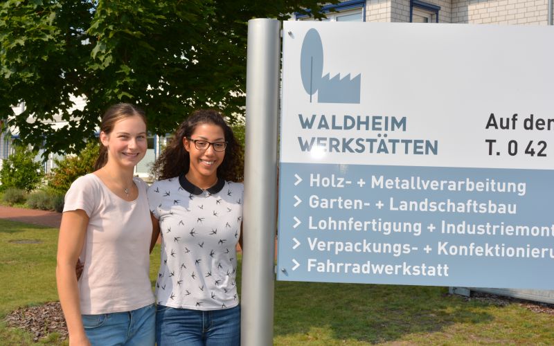 Zwei Mitarbeiterinnen vor einem Waldheimwerkstätten-Schild